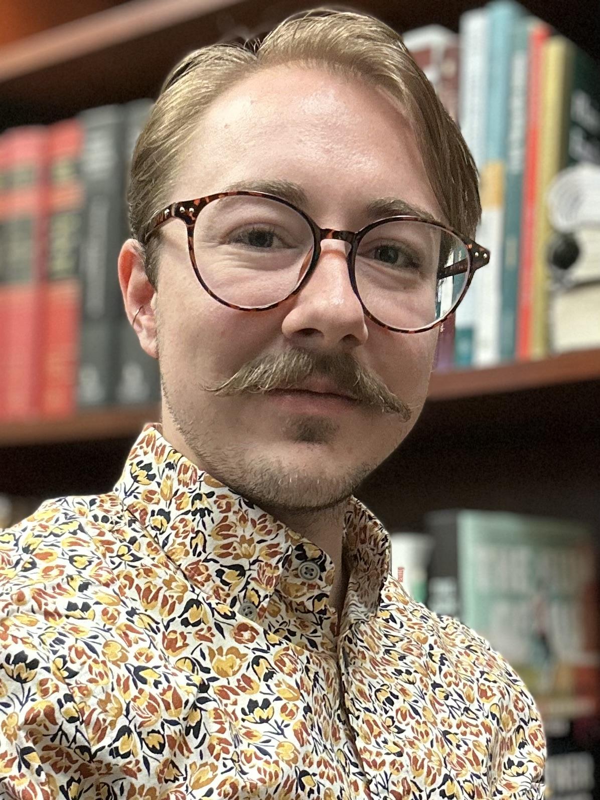 Young white trans guy with large glasses and a small handlebar mustache